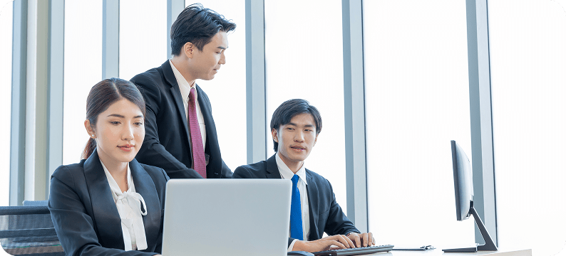 Three business professionals in formal attire working together at laptops and desktops in a modern office.