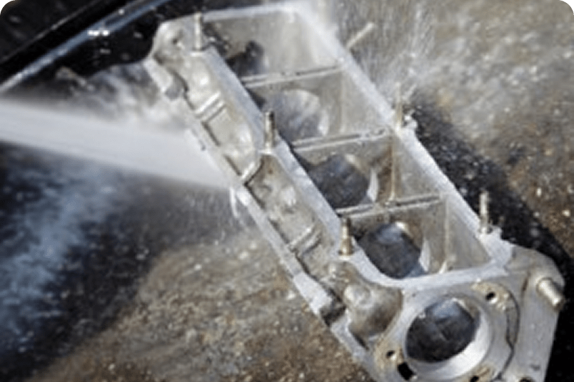 Aluminum engine part being cleaned with high-pressure water spray, showing surface preparation or industrial cleaning process.