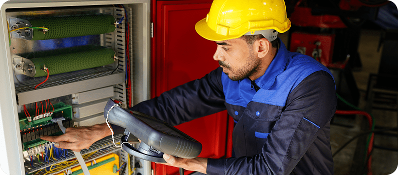 Technician in a yellow hard hat testing electrical control panel components with a handheld diagnostic device.