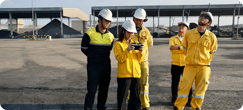 Group of industrial workers in safety gear operating a drone controller at an outdoor worksite.