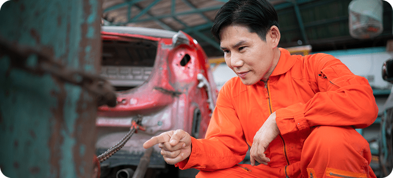 Man in orange coveralls inspecting chained metal near a damaged red car in an auto repair workshop.