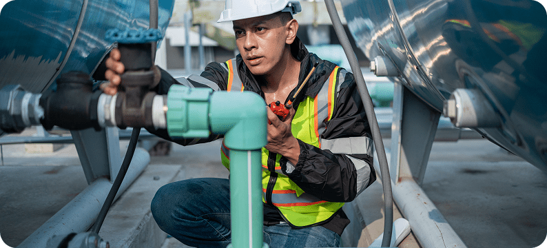 Engineer in safety gear inspecting and adjusting a pipeline valve between large industrial tanks.