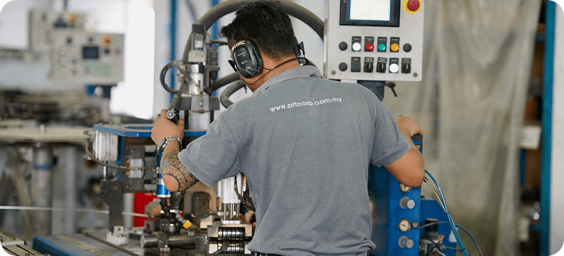 Technician operating a CNC machine with control panel in a manufacturing facility, wearing ear protection.