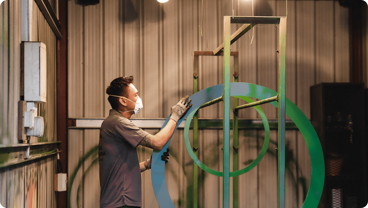 Worker wearing a mask spray-painting large circular gaskets in a metal fabrication workshop.